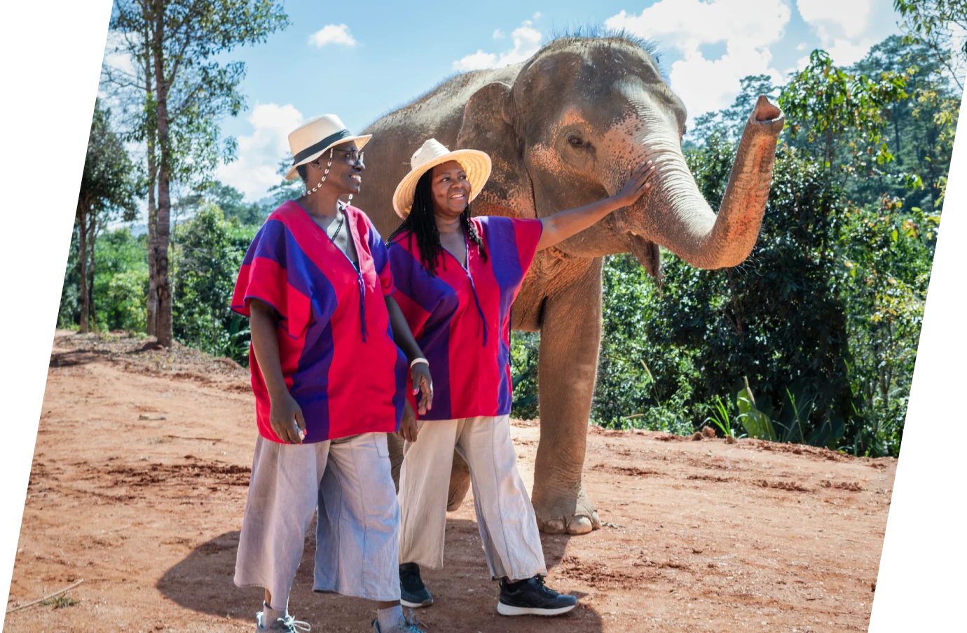 Two women in hats, walking with an elephant on vacation with travel insurance for Thailand