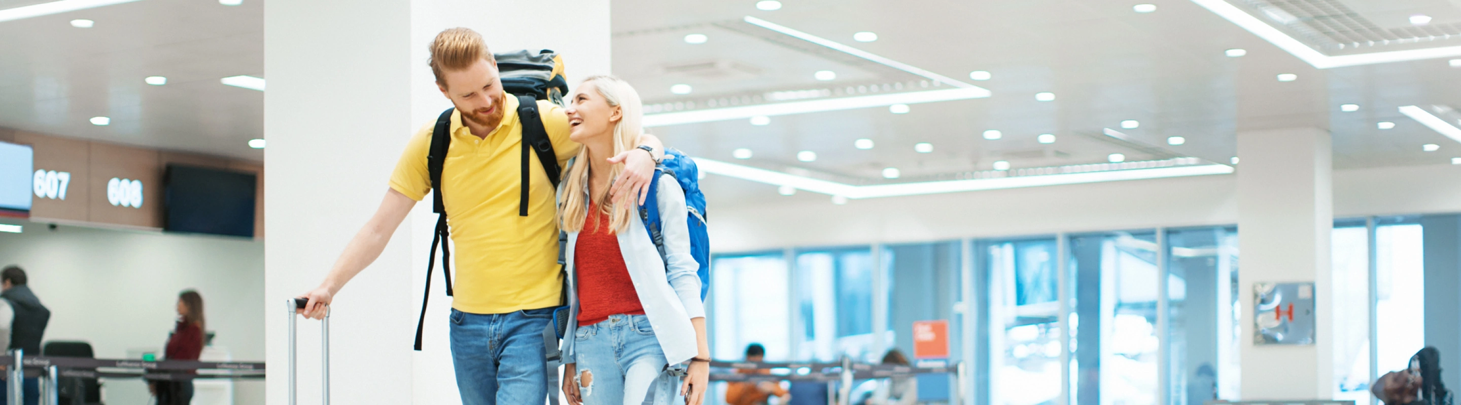 a young couple with cruise travel insurance and large backpacks walk alongside one another through an airport terminal 