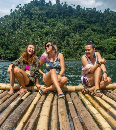 three young women sitting on a bamboo raft in the water, insured with travel insurance for the Philippines