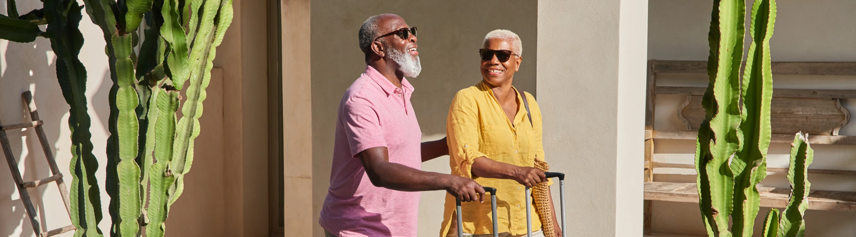 an older couple with travel insurance for wellness retreats rolls their suitcases through a desert garden with cacti