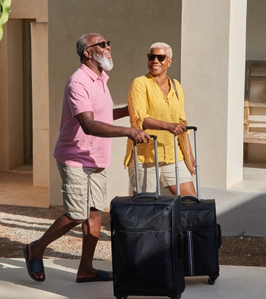 an older couple with travel insurance for wellness retreats rolls their suitcases through a desert garden with cacti