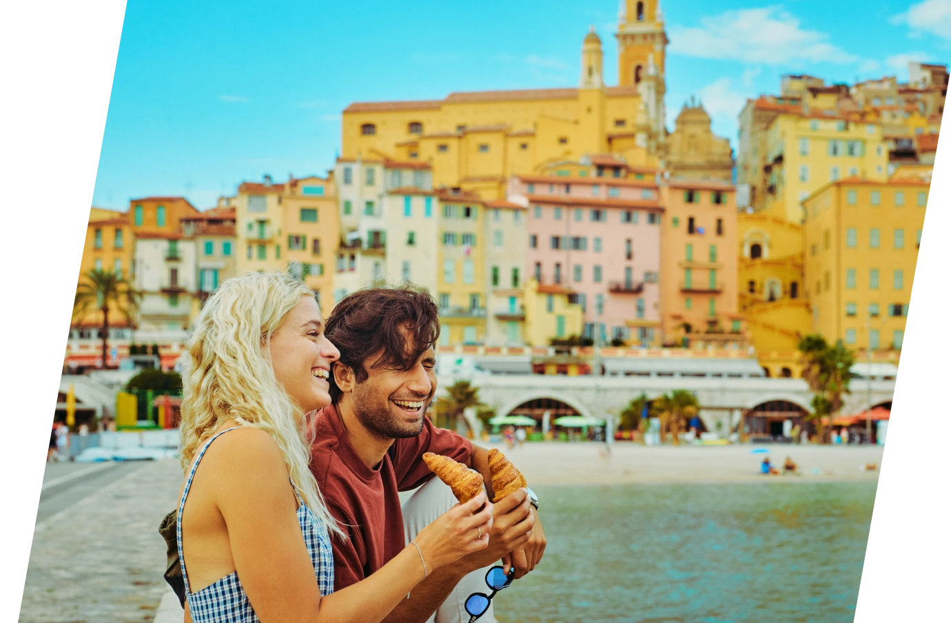 a young couple with travel insurance for culinary vacations eat croissants in a colorful Mediterranean seaside town 