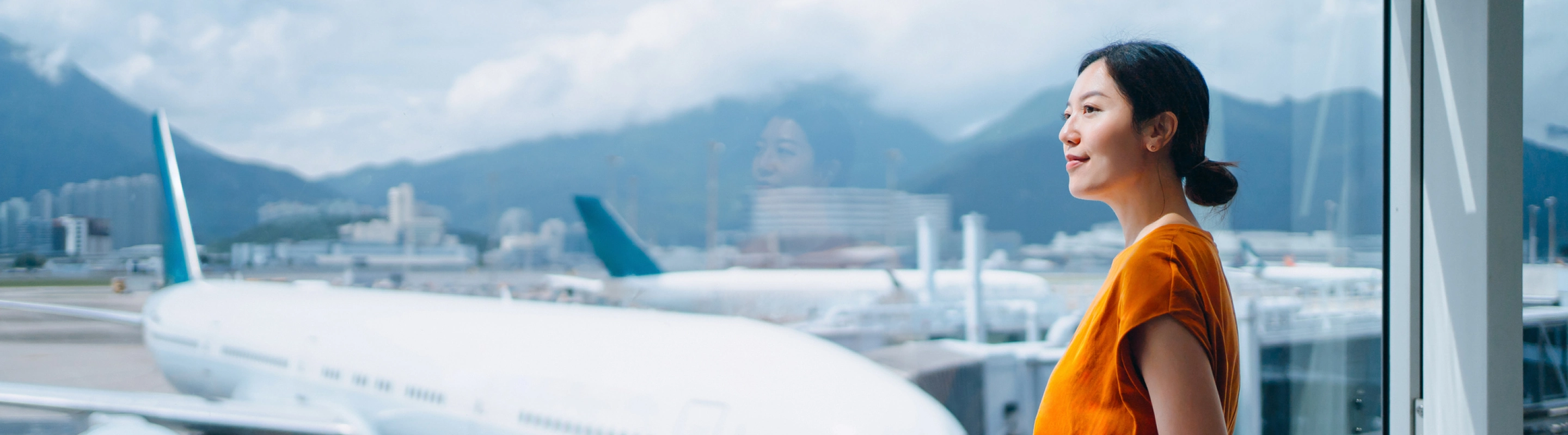 a young, Asian woman with travel insurance for nature trips stands by the window in an airport terminal to soak up views 