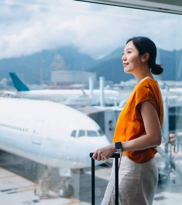 a young, Asian woman with travel insurance for nature trips stands by the window in an airport terminal to soak up views 