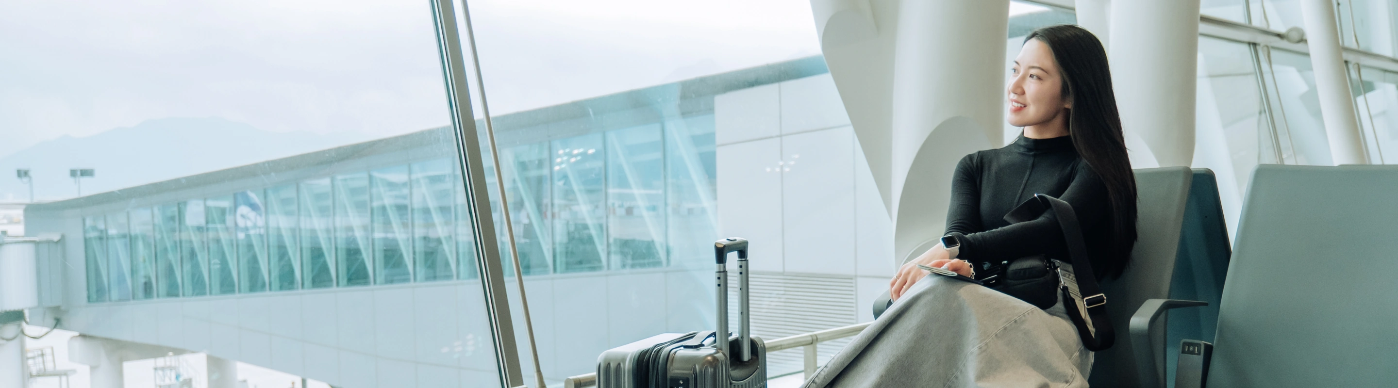 a young woman with pilgrimage travel insurance looks out a large window in an airport terminal with her suitcase beside her