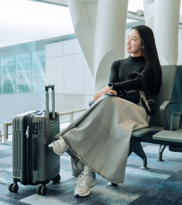 a young woman with pilgrimage travel insurance looks out a large window in an airport terminal with her suitcase beside her