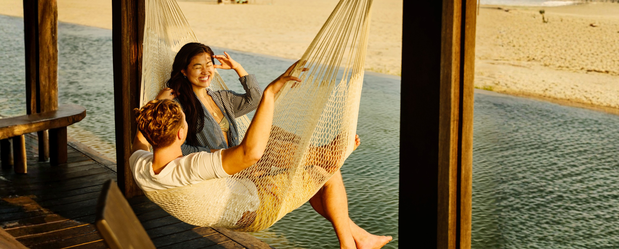 two young adults with travel insurance for wellness retreats sit on a hammock over a wooden deck by the ocean