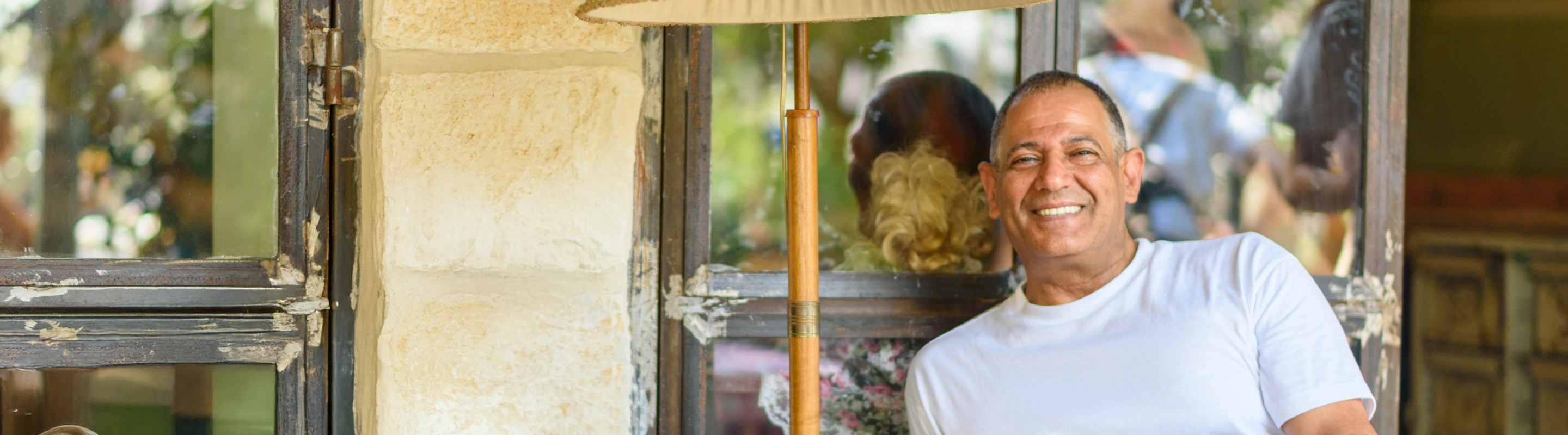 an older man with travel insurance for Israel smiles as he sits by a rustic window and traditional Israeli lamp 