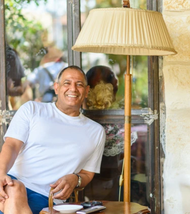 an older man with travel insurance for Israel smiles as he sits by a rustic window and traditional Israeli lamp 