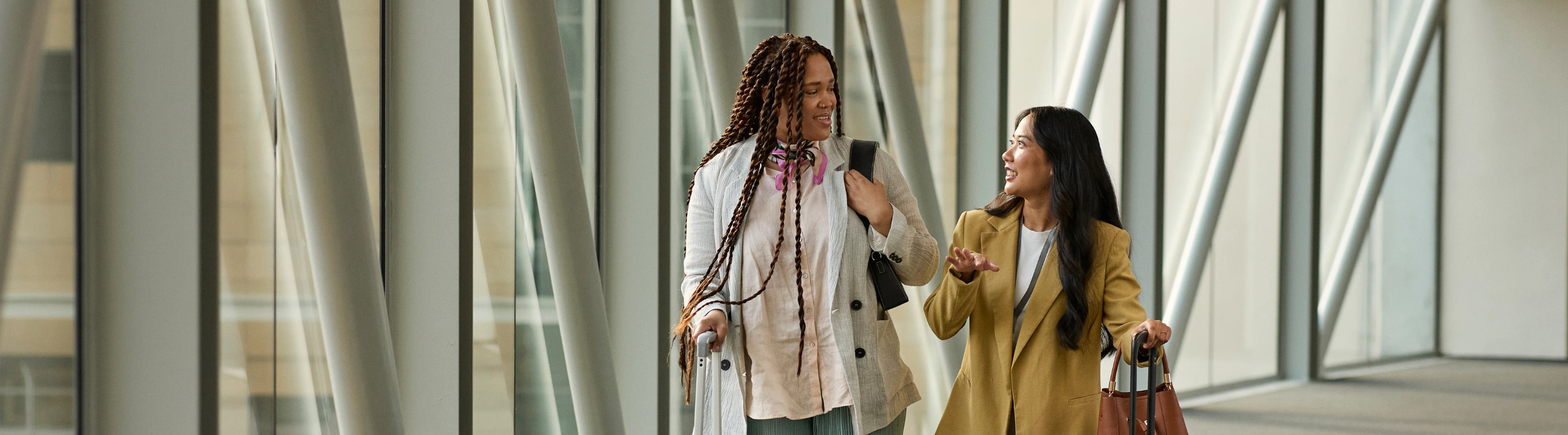 two middle-aged women with travel insurance for Israel walk down a modern glass corridor in an airport with their luggage