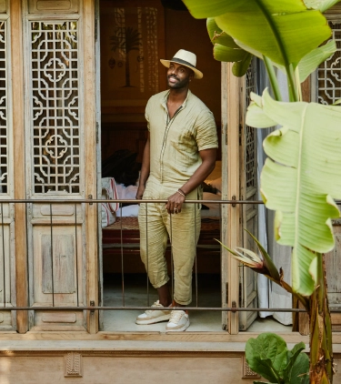 a young black man wearing a hat stands on a balcony outside his hotel room, insured with solo travel insurance 