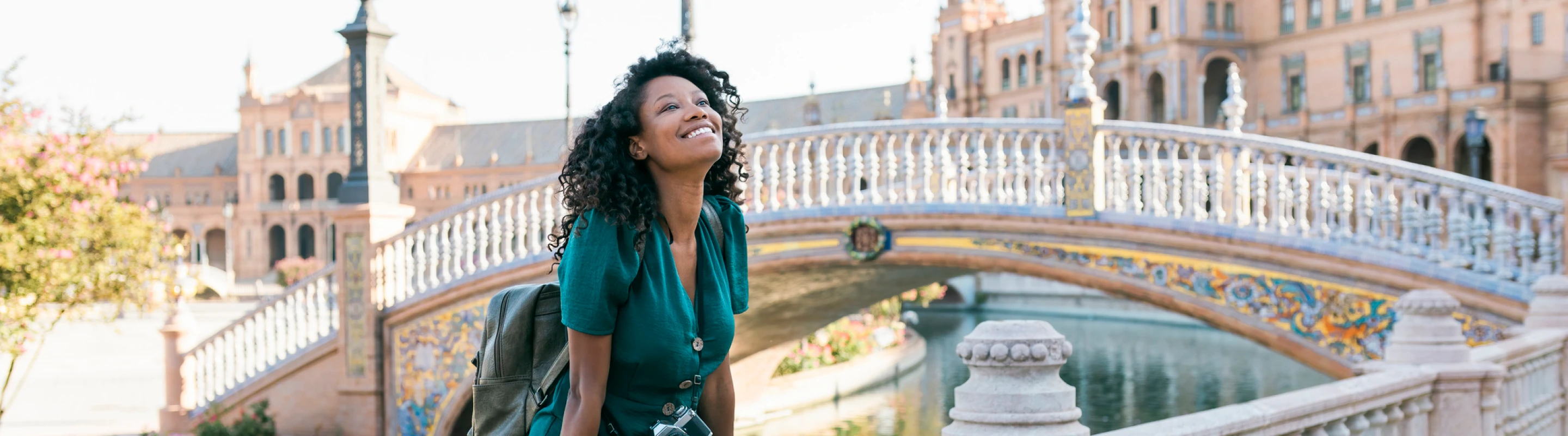 a young, mixed-race woman with a backpack and solo travel insurance sits near a decorative bridge and canal