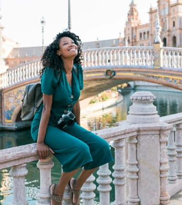 a young, mixed-race woman with a backpack and solo travel insurance sits near a decorative bridge and canal