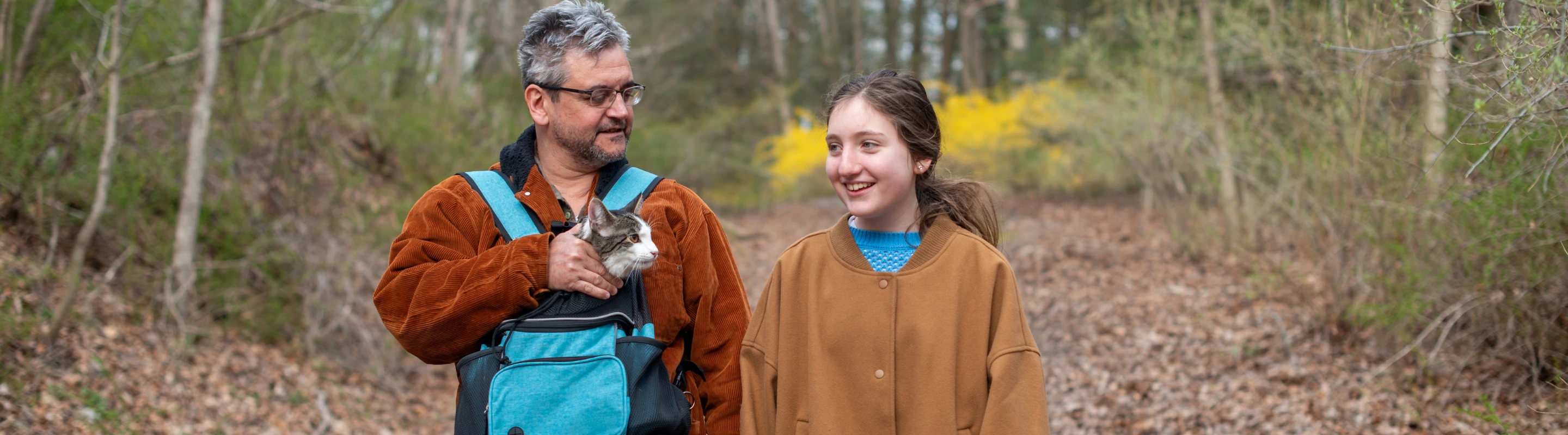 a father-daughter duo with pet travel insurance hike through a forest with their cat in a pouch 