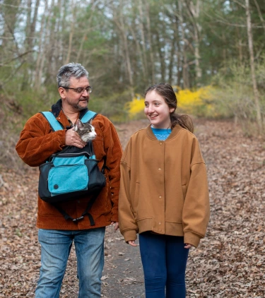 a father-daughter duo with pet travel insurance hike through a forest with their cat in a pouch 