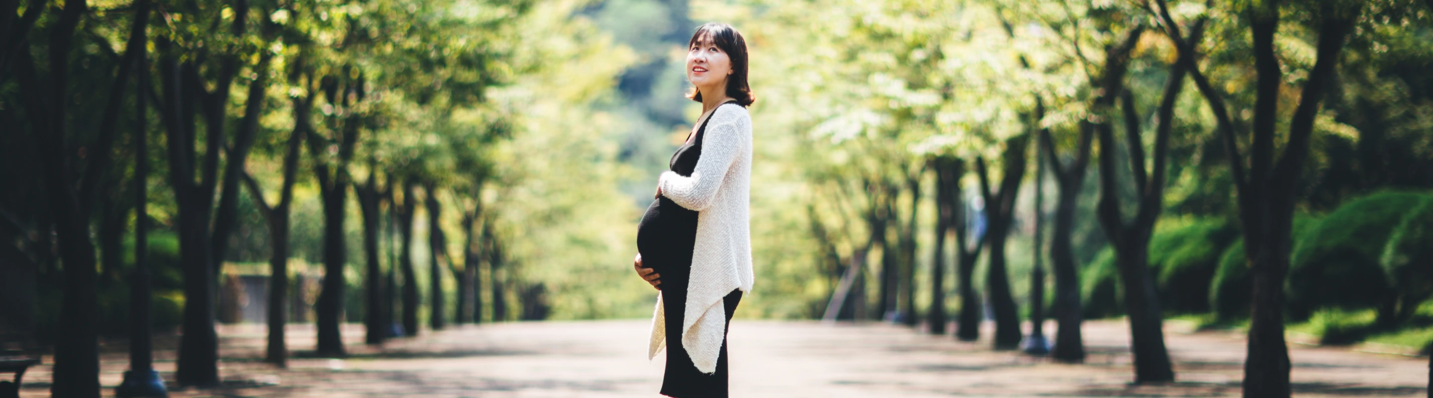 an Asian, pregnant woman with travel insurance for pregnancy stands in a park surrounded by green trees 