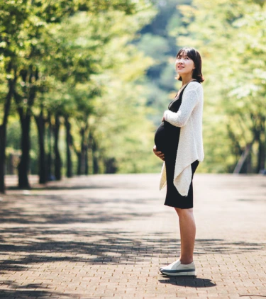 an Asian, pregnant woman with travel insurance for pregnancy stands in a park surrounded by green trees 