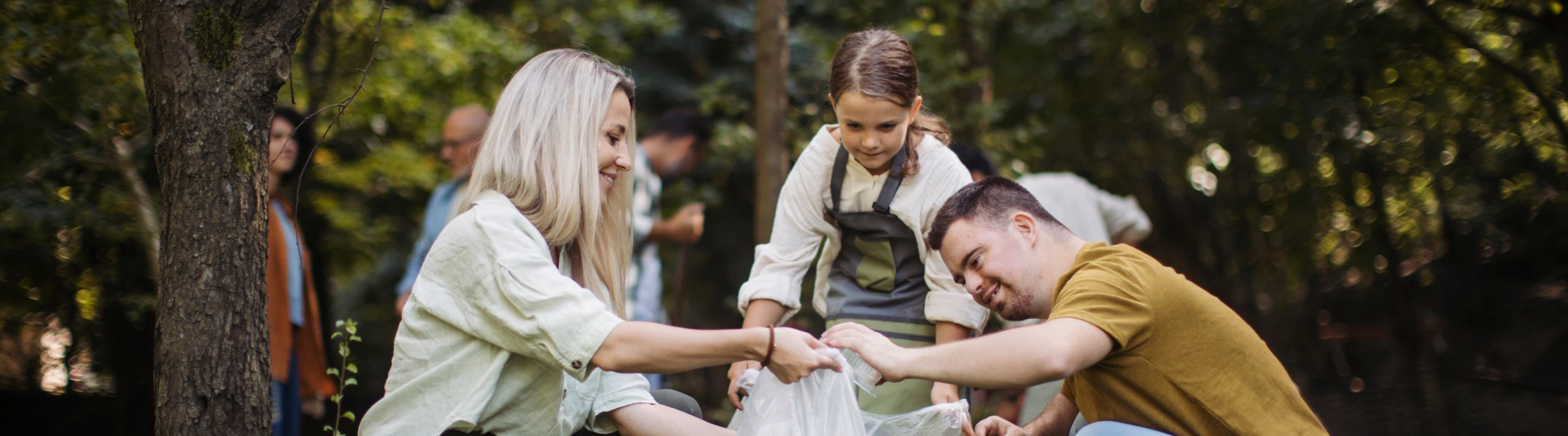 Two adults with volunteer travel insurance and a young girl putting trash into a trash bag in a park