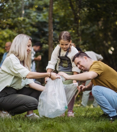 Two adults with volunteer travel insurance and a young girl putting trash into a trash bag in a park