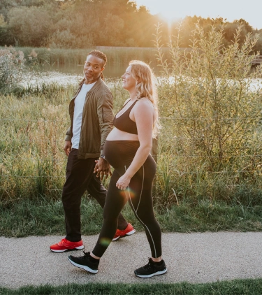 a pregnant, mixed-race couple with travel insurance for pregnancy walks through a green park at sunset
