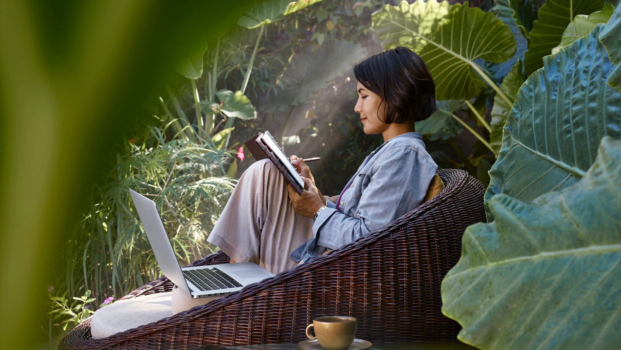 a woman sits in a garden near her laptop as she writes down travel insurance claims tips in her notebook
