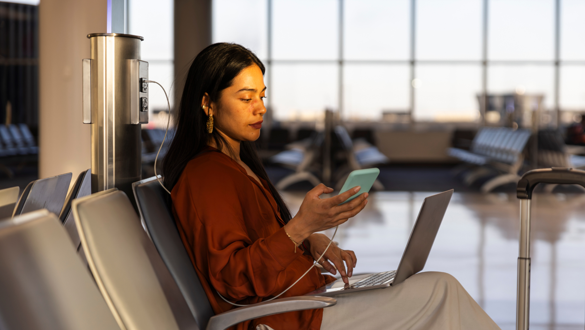 a woman looks at her phone and holds her laptop near a charging station in an airport after reading our data privacy tips