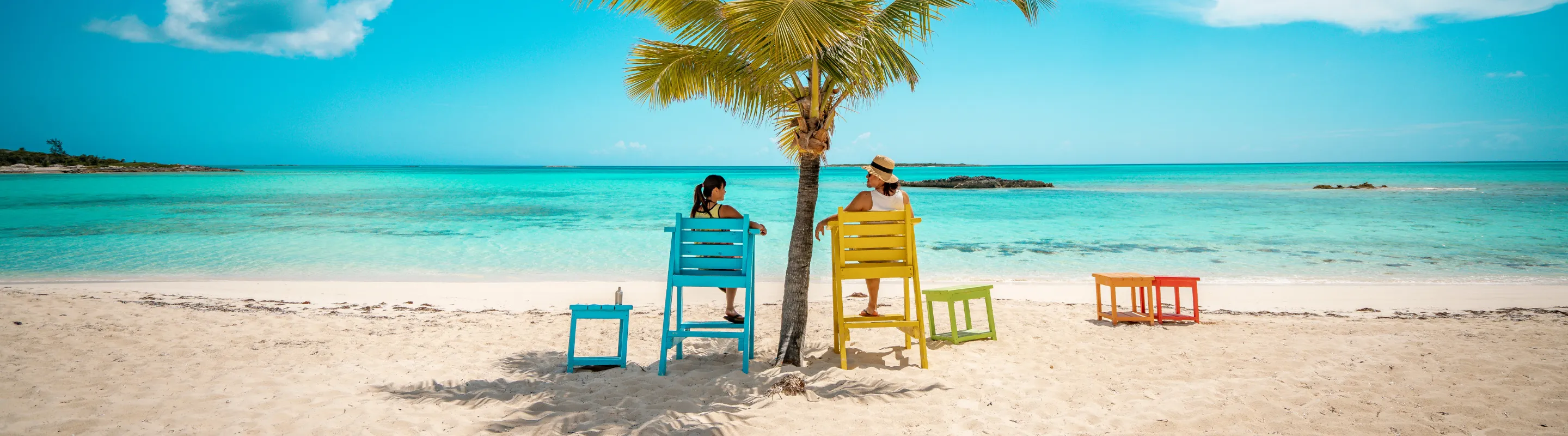 two women with travel insurance for the Bahamas relax in colorful chairs beneath a palm tree on a white-sand beach