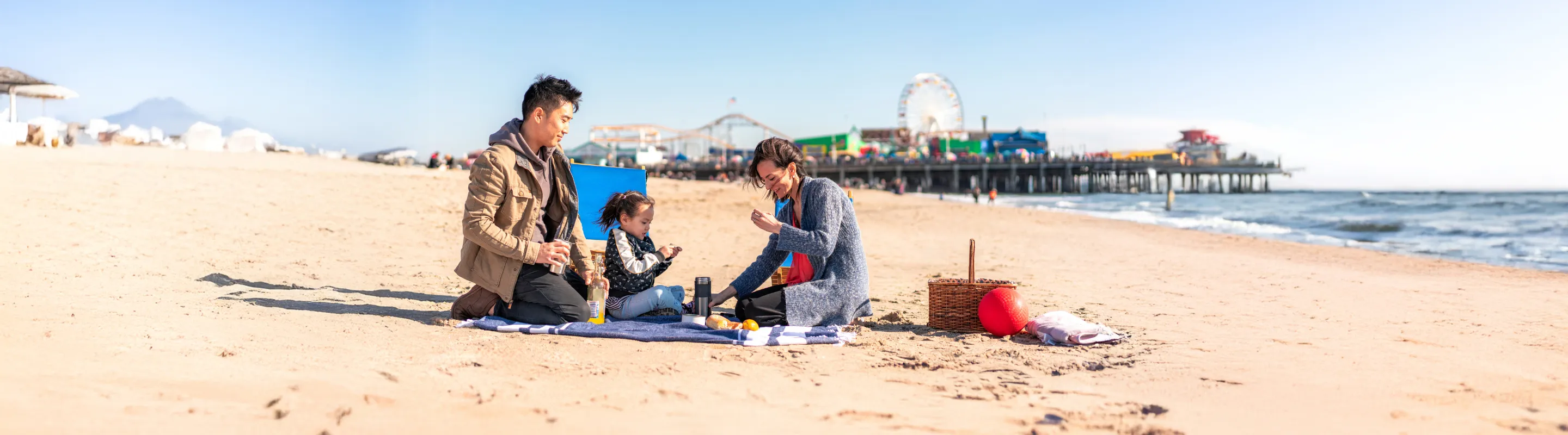 two parents, with travel insurance for California, and their toddler enjoy a picnic on a beach near Santa Monica Pier
