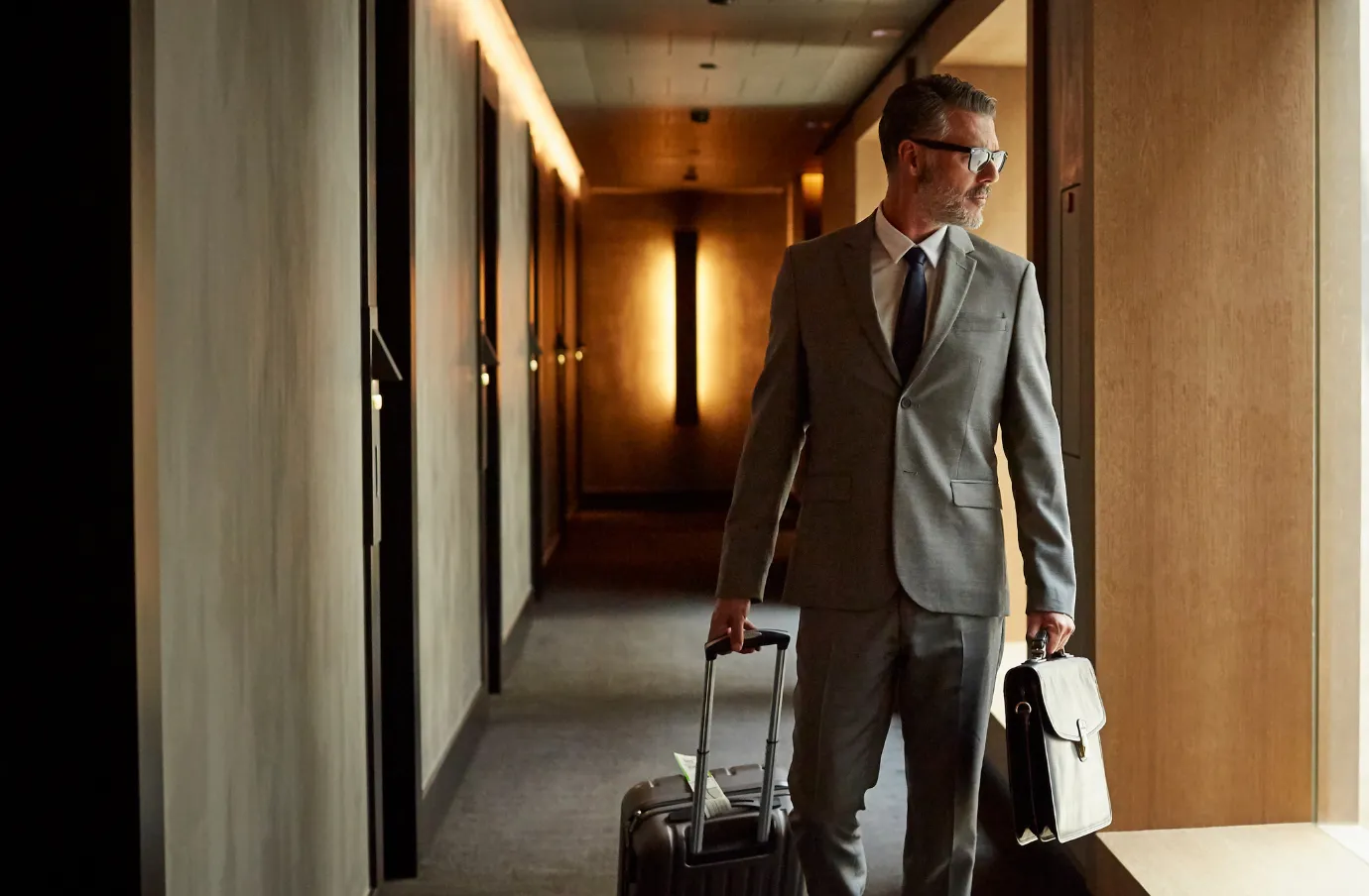 a businessman with travel insurance for Washington, D.C. walks through a hotel corridor with a rolling suitcase