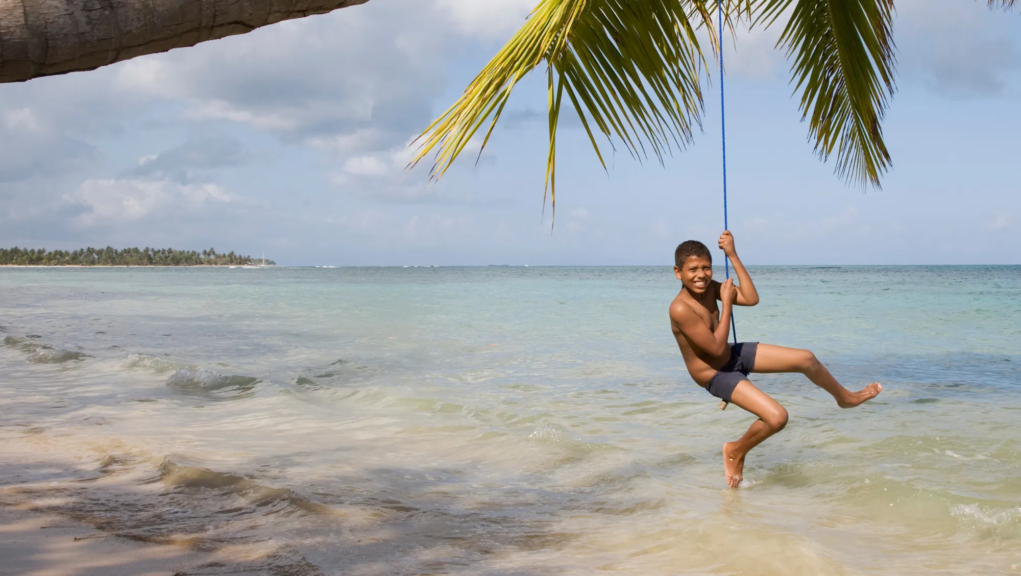 a boy with travel insurance for hurricanes plays on a rope swing over clear coastal water beneath a palm tree