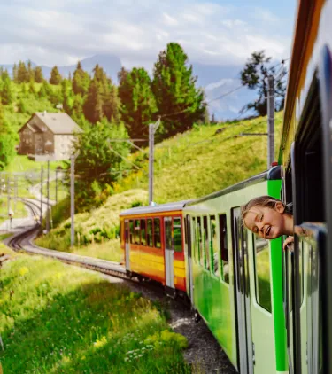 a girl pokes her head out of the window of a colorful train in the Swiss Alps, insured with travel insurance for Switzerland 