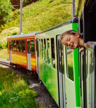 a girl pokes her head out of the window of a colorful train in the Swiss Alps, insured with travel insurance for Switzerland 