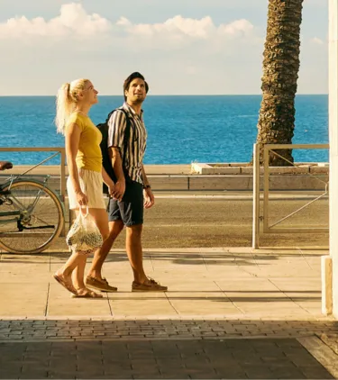 a young couple with travel insurance for beach trips holds hands as they walk palm trees and the ocean 