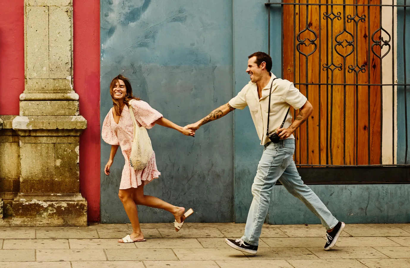 couple holds hands while walking past colorful colonial buildings in Mexico