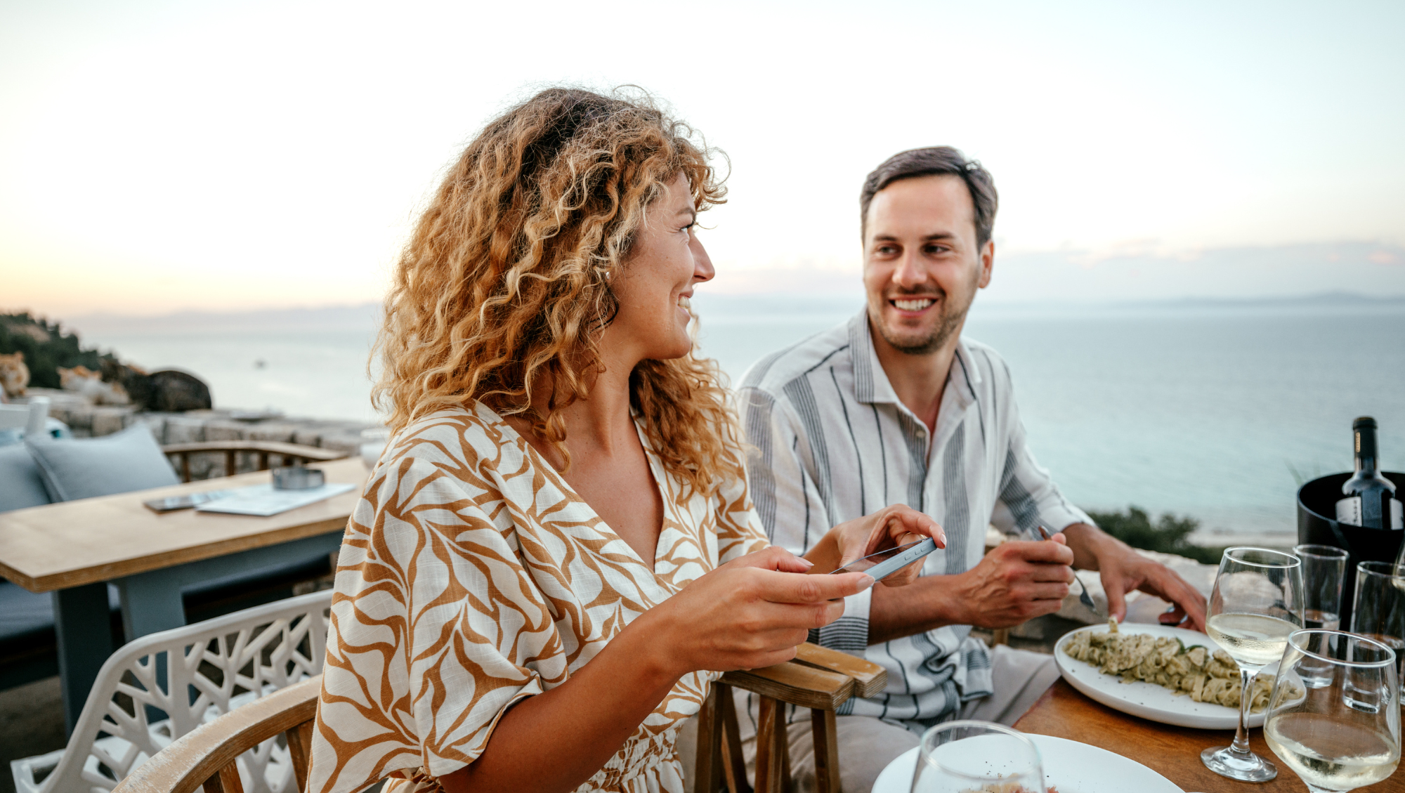 a couple dines at a seaside restaurant, and the woman checks the travel app on her phone 