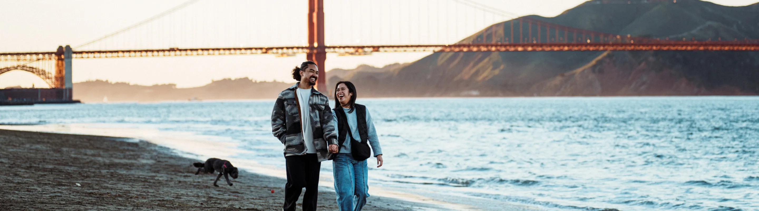 a couple with domestic travel insurance walk along San Francisco's shoreline with the Golden Gate Bridge in the background
