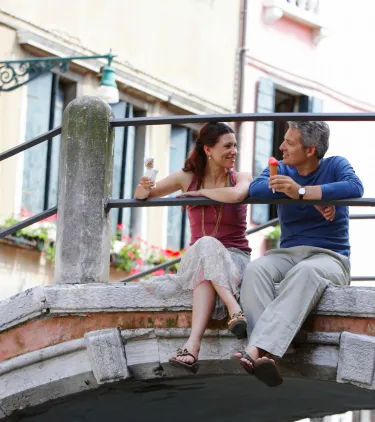 a middle-aged couple sits on a bridge and eats gelato in Venice, insured with travel insurance for Italy