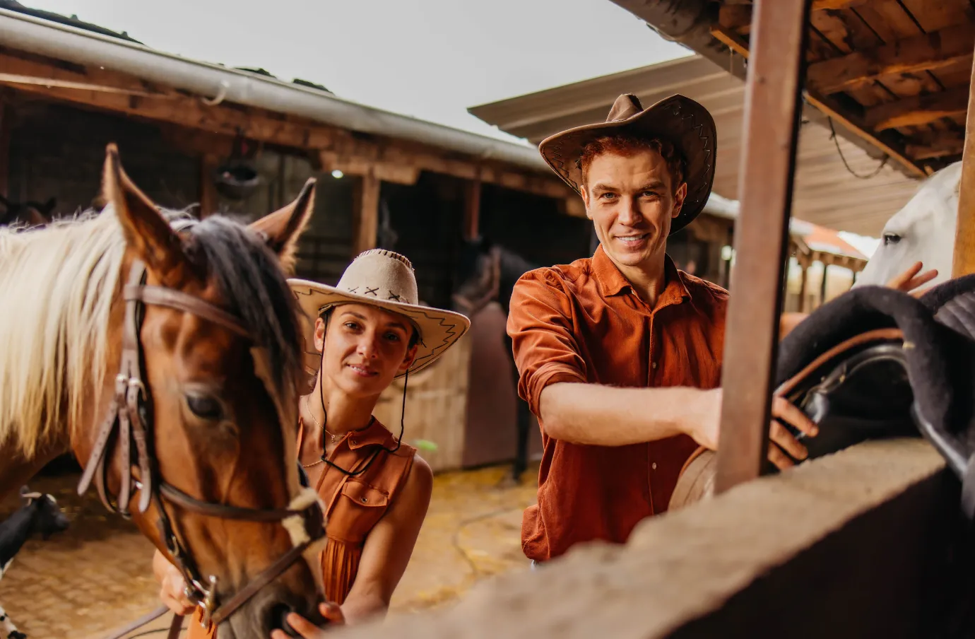 a young couple with cowboy hats and travel insurance for Texas pose by horses at a ranch