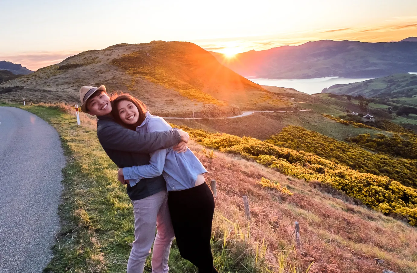 a young couple with travel insurance for New Zealand hugs on the side of a winding mountain road at sunset 