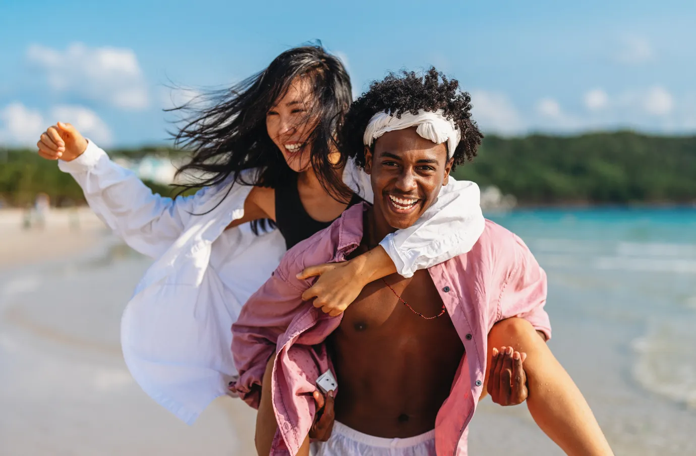 a smiling couple with travel insurance for the Bahamas enjoys a piggyback ride along a tropical beach