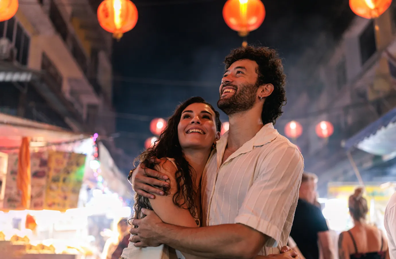 a couple with travel insurance for Thailand embraces beneath red lanterns in a lively night market
