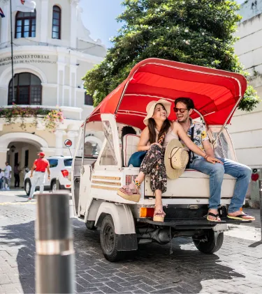 a couple with travel insurance for the Dominican Republic explore a cultural district while riding on a tour cart 