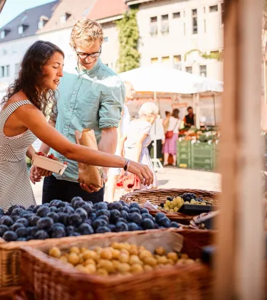 a young couple shops at a local market for fruit, insured with travel insurance for Germany