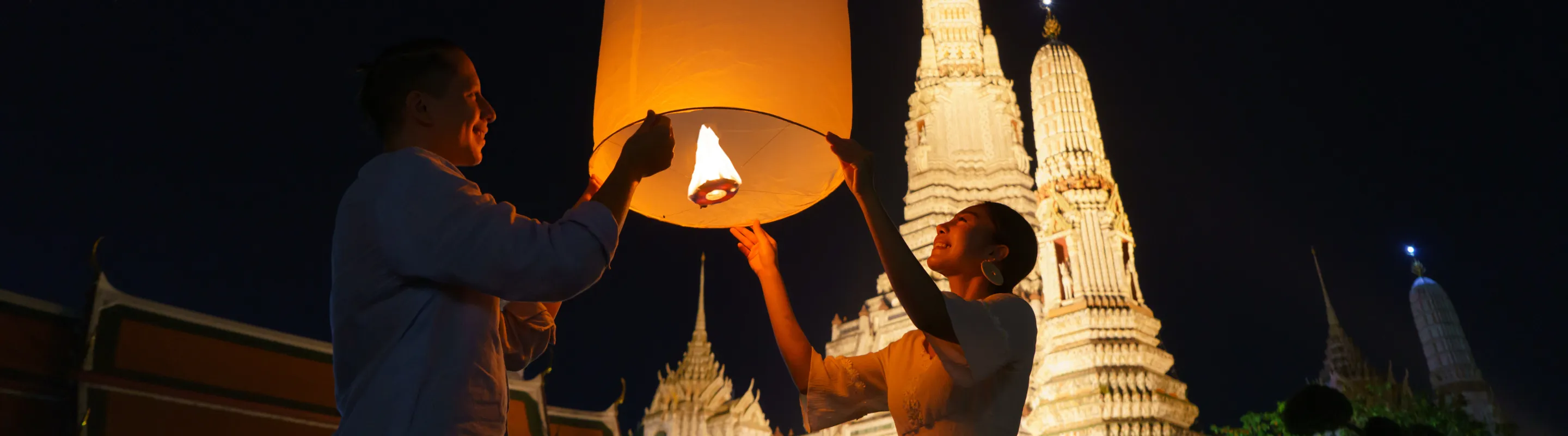 a young couple with travel insurance for Thailand releases a lantern into the sky near Wat Arun temple 