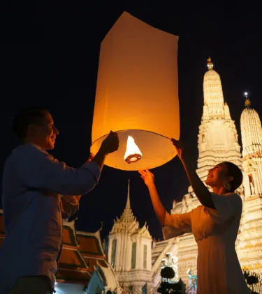 a young couple with travel insurance for Thailand releases a lantern into the sky near Wat Arun temple 