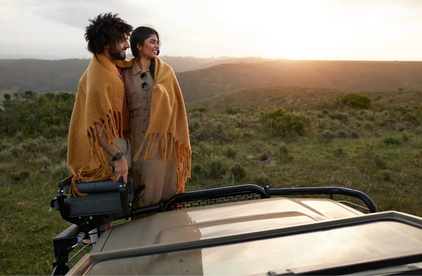 a couple stands beside a safari vehicle at sunset, wrapped in blankets and insured with travel insurance for Africa 