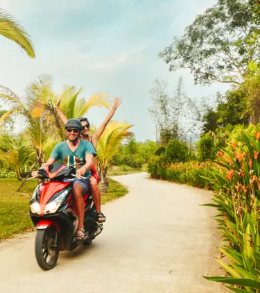 a couple with travel insurance for Vietnam rides a red scooter along a winding road lined with palm trees