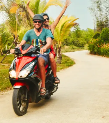 a couple with travel insurance for Vietnam rides a red scooter along a winding road lined with palm trees