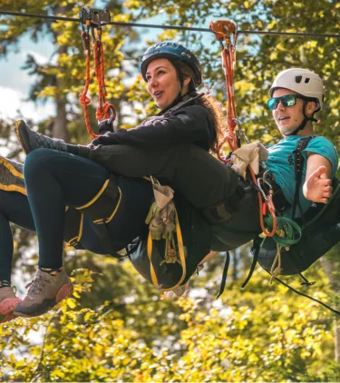 a young couple with adventure travel insurance tandem zip line through treetops on a blue-sky day