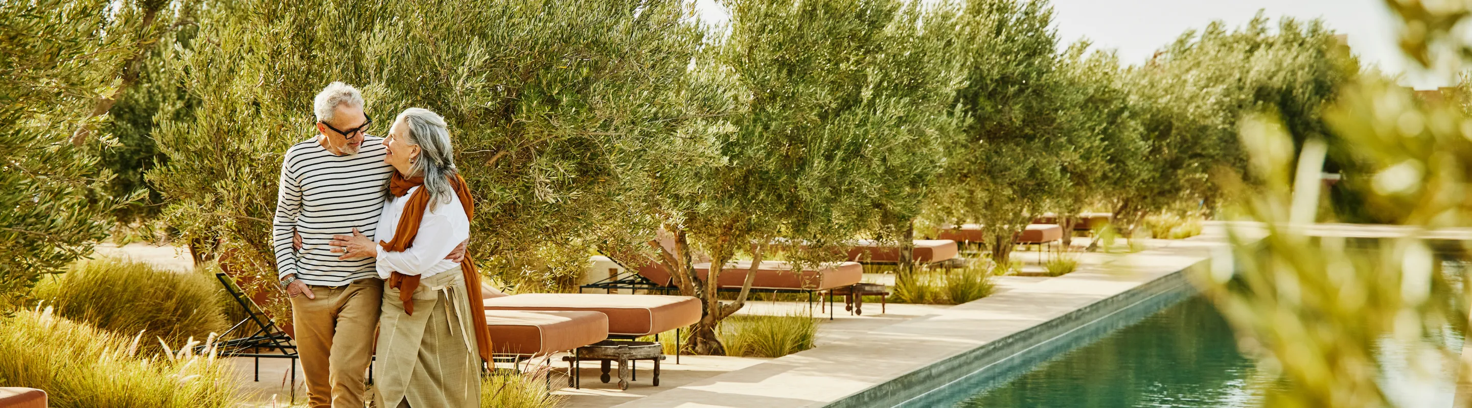 an elderly couple with long-term travel insurance walks through a garden-lined poolside resort 